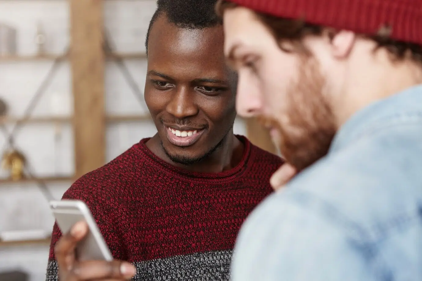 Un joven hombre negro, entusiasmado y feliz, con un suéter, le muestra a su amigo blanco a la moda una cuenta de blog inspiradora en las redes sociales. Dos mejores amigos varones usan un teléfono móvil juntos en un café. Enfoque selectivo.